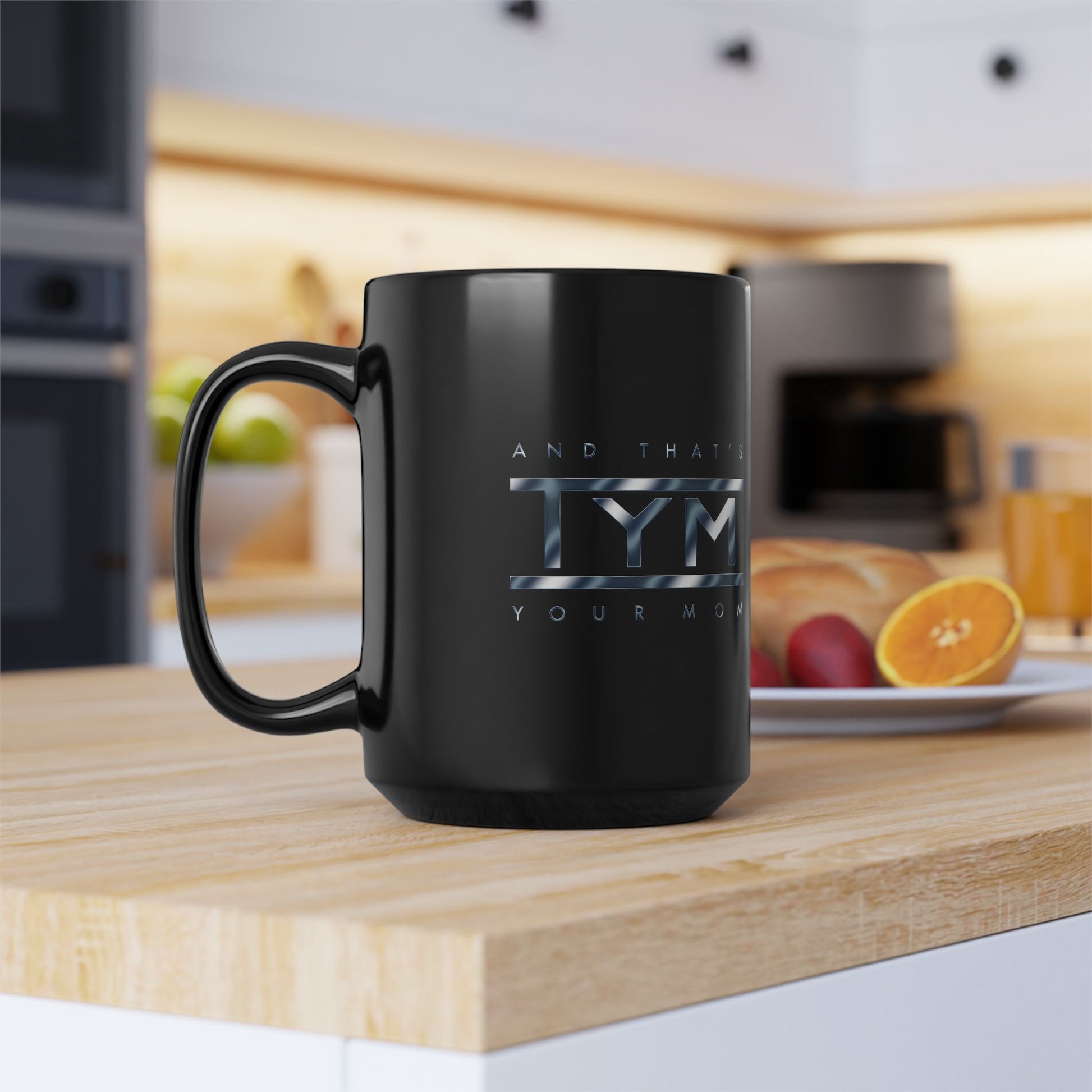 black coffee mug featuring TYM and that's your mom chrome blue design on a kitchen counter next to fruit